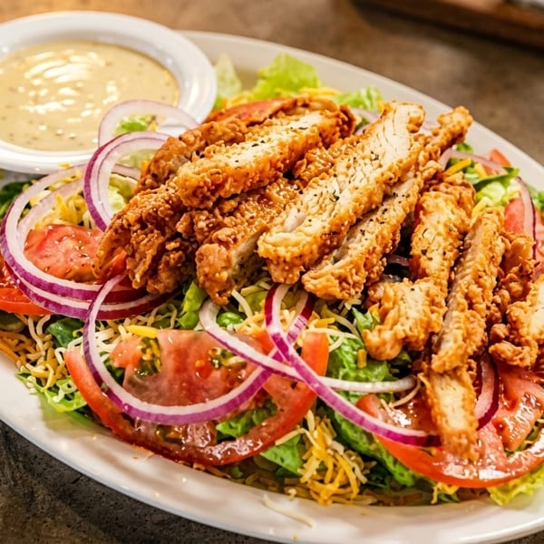 Crispy fried chicken salad with lettuce, tomatoes, red onions, shredded cheese, and a side of dressing.