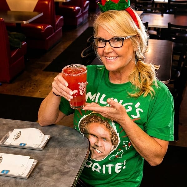 Blonde woman in elf headband and Buddy the Elf shirt holding a red cocktail at a diner counter.